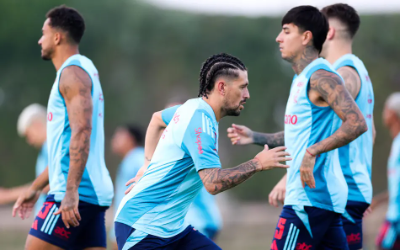 Flamengo players in light blue and white training kits practicing drills on a grass pitch, passing and running while focused and determined; blurred trees and training ground equipment visible in the background